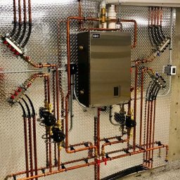 Termo-Serwis Ireneusz Radecki - Nowoczesny, kompaktowy piec grzewczy IBC z miedzianą instalacją i rozdzielaczami na tle ściany z blachy ryflowanej.