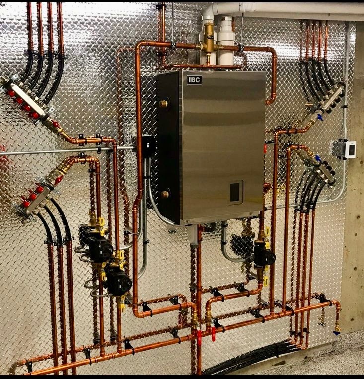 Nowoczesny, kompaktowy piec grzewczy IBC z miedzianą instalacją i rozdzielaczami na tle ściany z blachy ryflowanej.