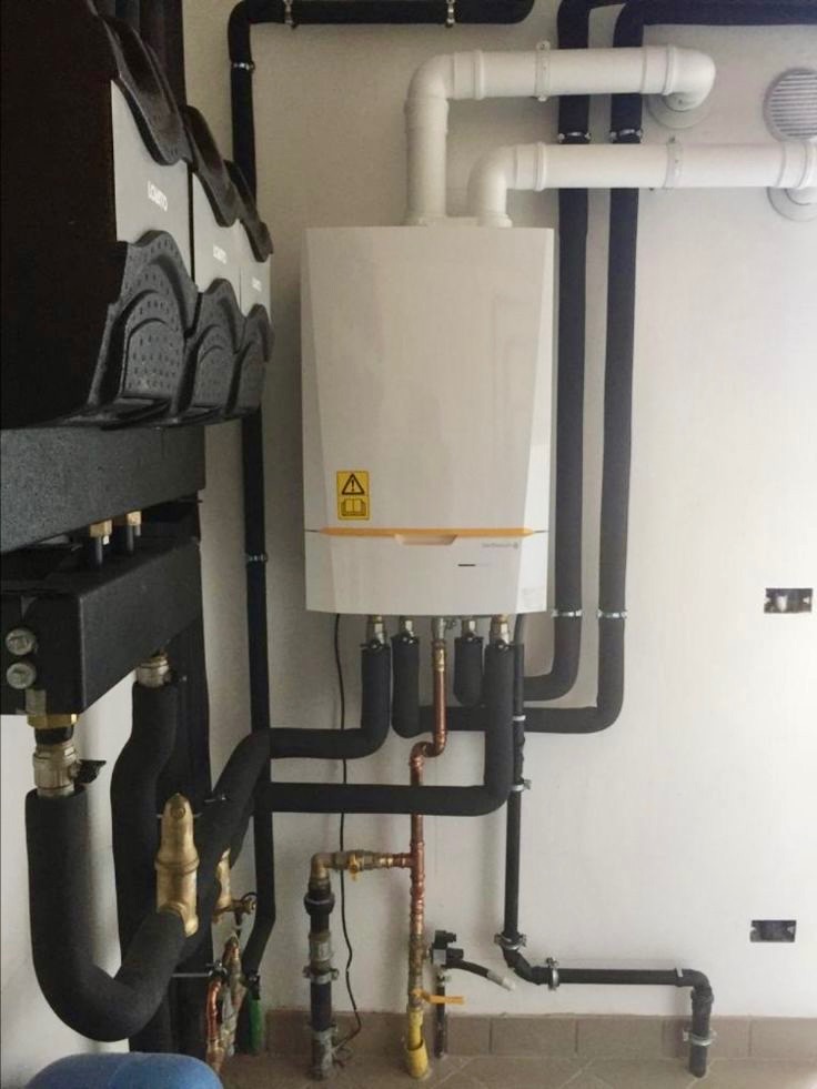 Nowoczesny, biały piec gazowy z żółtymi akcentami, podłączony do systemu rur w izolacji, zamontowany na białej ścianie z widocznymi przyłączami i elementami instalacji hydraulicznej.