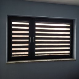 Maxus Jakub Maj - Ciemna rama okienna z roletami dzień noc w poziome pasy, zamontowana na tle błękitnej ściany z szarawym parapetem.