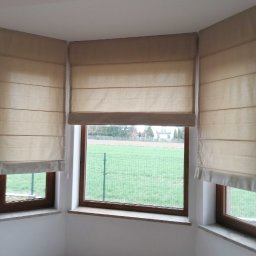 Maxus Jakub Maj - Beżowe plisy rzymskie zamontowane w trzech oknach tworzących wykusz, widok na zielony trawnik i dom w tle.