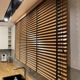 Maxus Jakub Maj - Drewniane żaluzje poziome z czarnymi sznurkami w kuchni, widoczne zlew, bateria i oświetlenie podszafkowe.