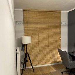 Maxus Jakub Maj - Drewniane żaluzje poziome w ciepłym odcieniu, zamontowane w oknie, widoczne z perspektywy wnętrza pokoju z lampą na trójnogu i fragmentem biurka z krzesłem.