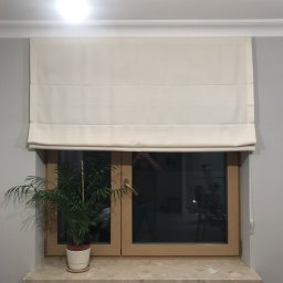 Maxus Jakub Maj - Kremowa roleta rzymska zamontowana w oknie z drewnianą ramą, widok z wnętrza mieszkania w Świeciu, na parapecie doniczka z zieloną rośliną.