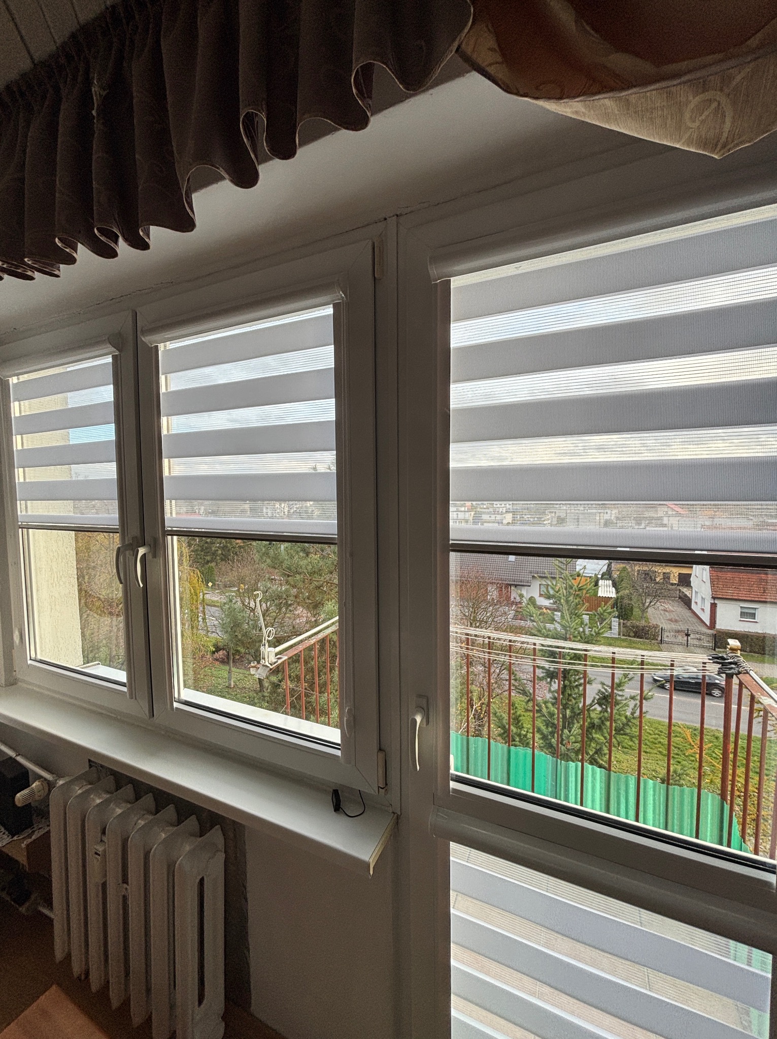 Trzyczęściowe okno z zamontowanymi roletami dzień i noc z widokiem na balkon i ulicę z zielonym ogrodzeniem.