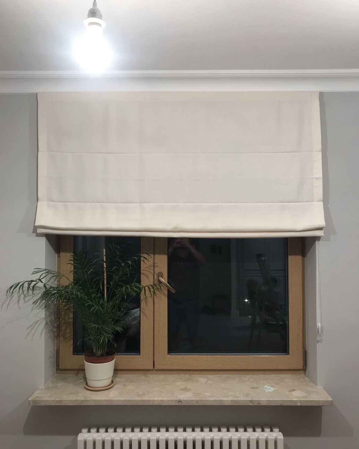 Kremowa roleta rzymska zamontowana w oknie z drewnianą ramą, widok z wnętrza mieszkania w Świeciu, na parapecie doniczka z zieloną rośliną.