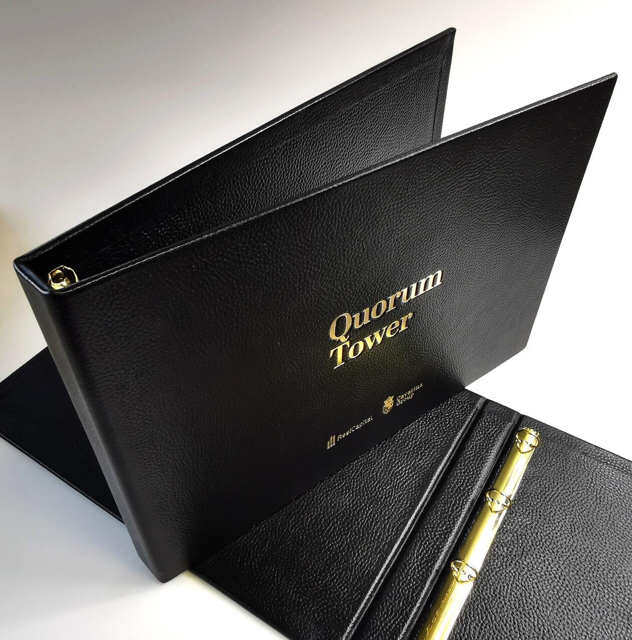 Czarny, skórzany segregator A3 z tłoczonym złotym napisem 'Quorum Tower' i logotypami, widoczne złote ringi.