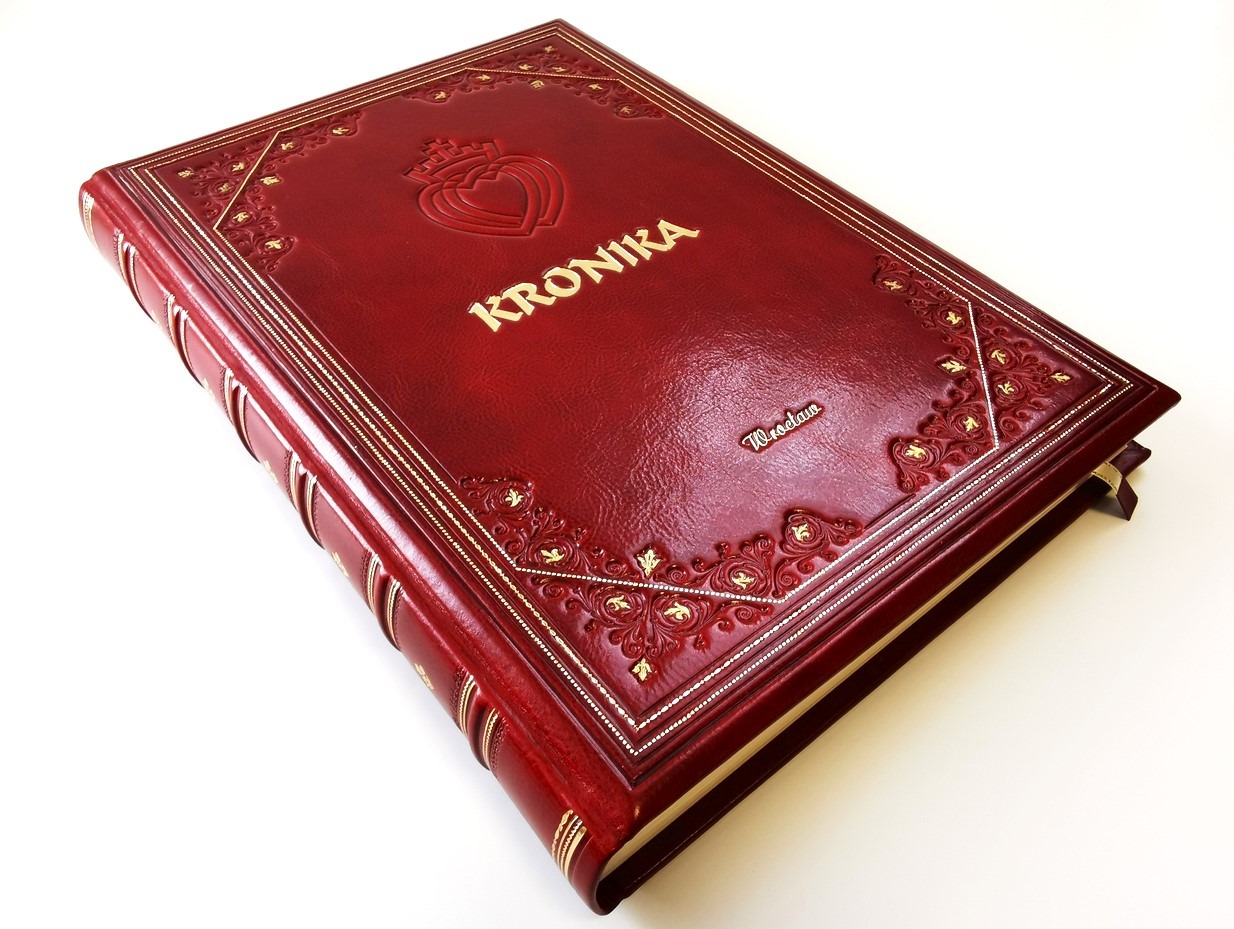 Oprawiona w czerwoną skórę kronika kościelna z tłoczeniami, złoceniami i napisem 'Kronika' w kolorze złotym, szyta ręcznie, z widocznym formatem A3 i grubością bloku.