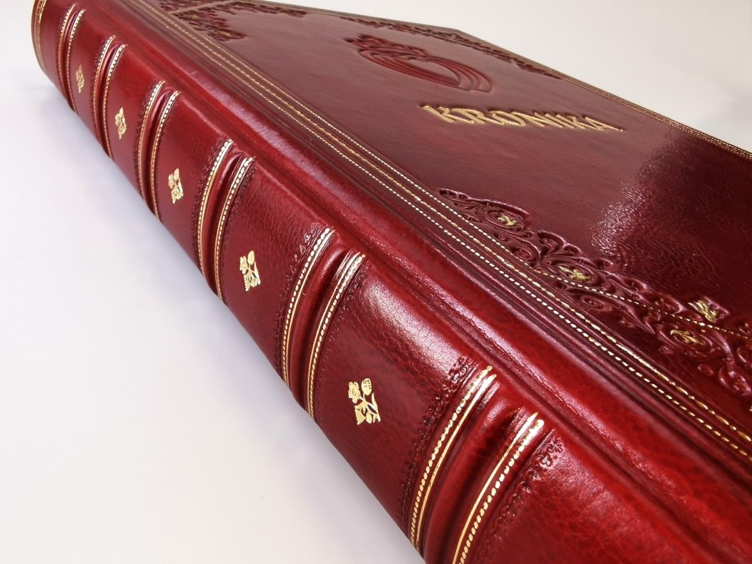 Oprawiona w czerwoną skórę kronika formatu A3 z tłoczonym złotym napisem i zdobieniami, widoczny grzbiet z wypukłymi żebrami i złotymi paskami.