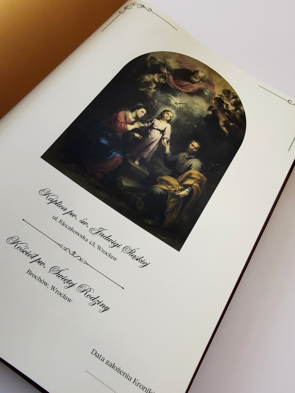 Otwarta księga pamiątkowa w formacie A3 z ilustracją religijną i tekstem o kościele, elegancka czcionka, ozdobne winiety w rogach strony, miejsce na wpisanie daty założenia kroniki.