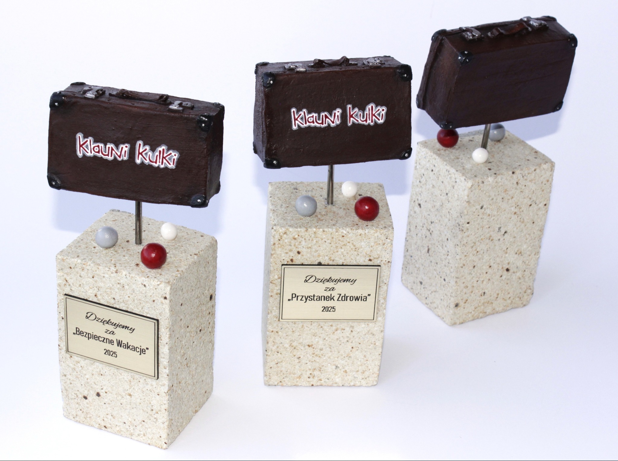 Trzy statuetki z motywem walizki, każda na kamiennej podstawie z grawerowaną tabliczką z podziękowaniami na rok 2025, z akcentami w postaci kulek.