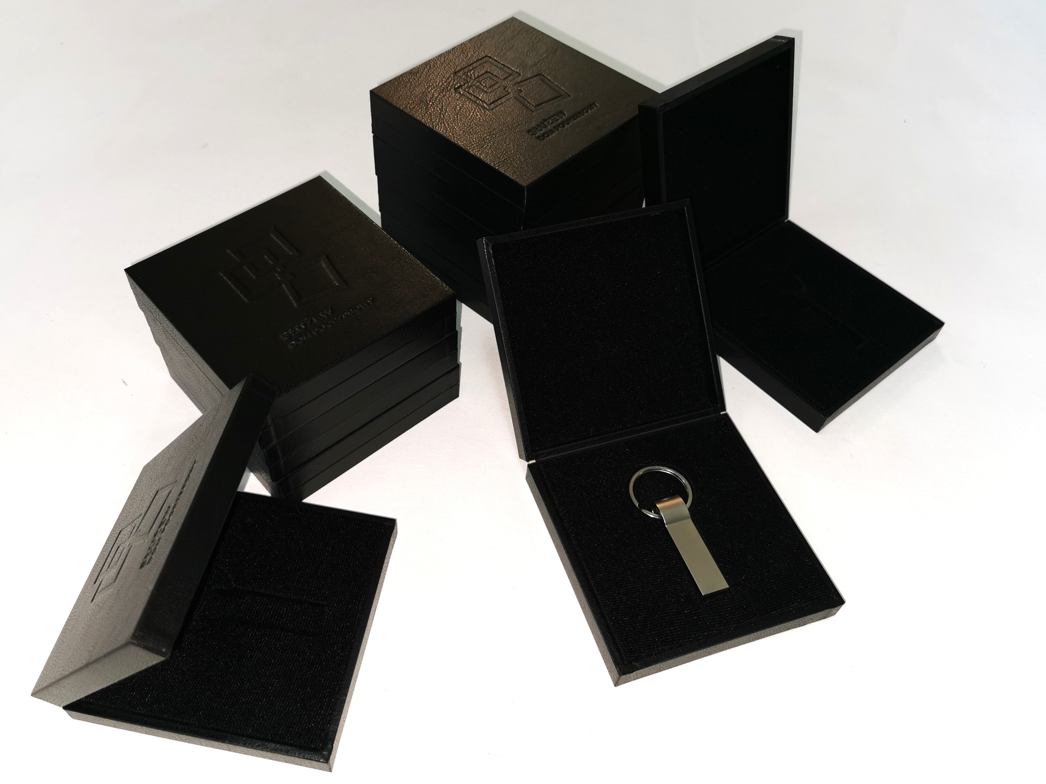 Czarne, kwadratowe pudełka z tłoczonym logo, wyściełane welurem, przeznaczone na pendrive. Jedno pudełko otwarte, w środku pendrive. Eleganckie opakowanie.