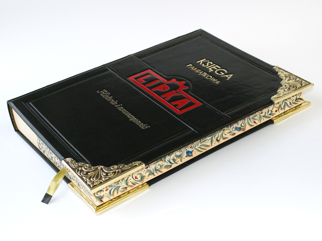 Elegancka, czarna księga pamiątkowa w skórzanej oprawie, zdobiona mosiężnymi narożnikami i ręcznie malowanymi brzegami bloku. Widoczne tłoczenia w kolorze czerwonym i złotym.