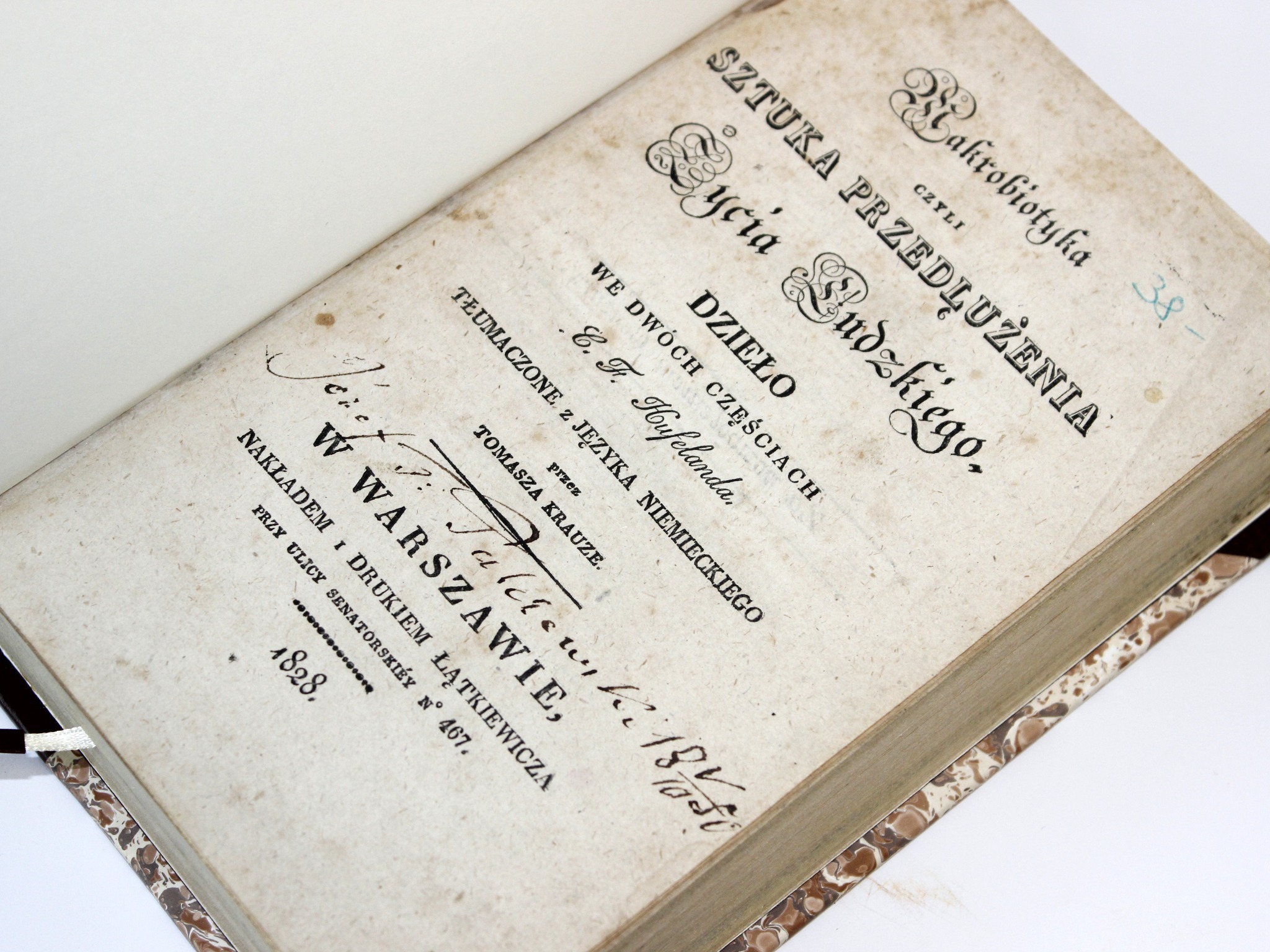 Otwarta, stara książka z 1828 roku z tytułem 'Sztuka przedłużenia życia ludzkiego' w ozdobnej czcionce, leżąca na białym tle. Widoczna faktura papieru i zdobiona okładka.