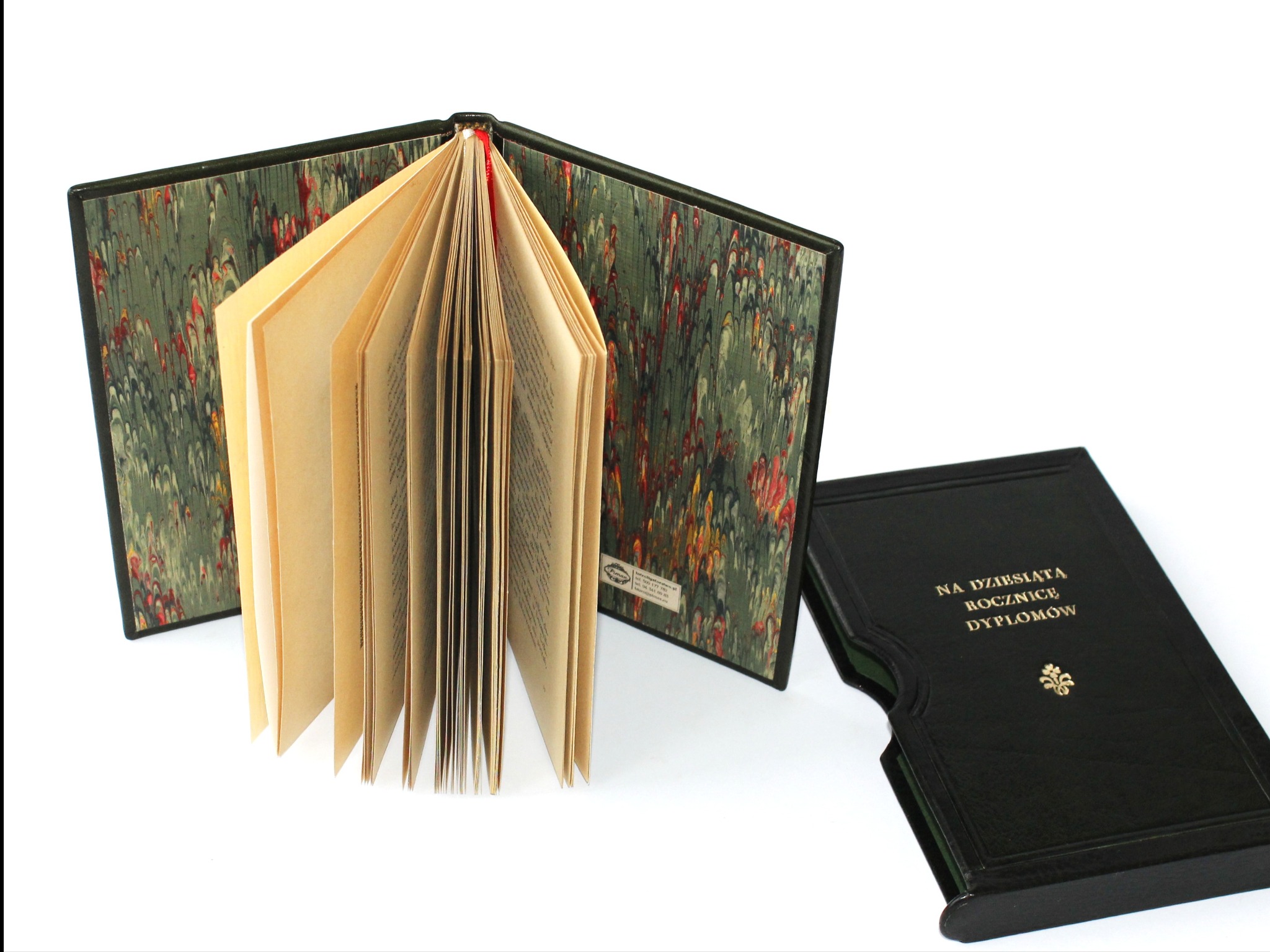 Otwarta książka w skórzanej oprawie z marmurkowym wzorem, obok etui z napisem 'Na dziesiątą rocznicę dyplomów', na białym tle. Introligatorska robota.