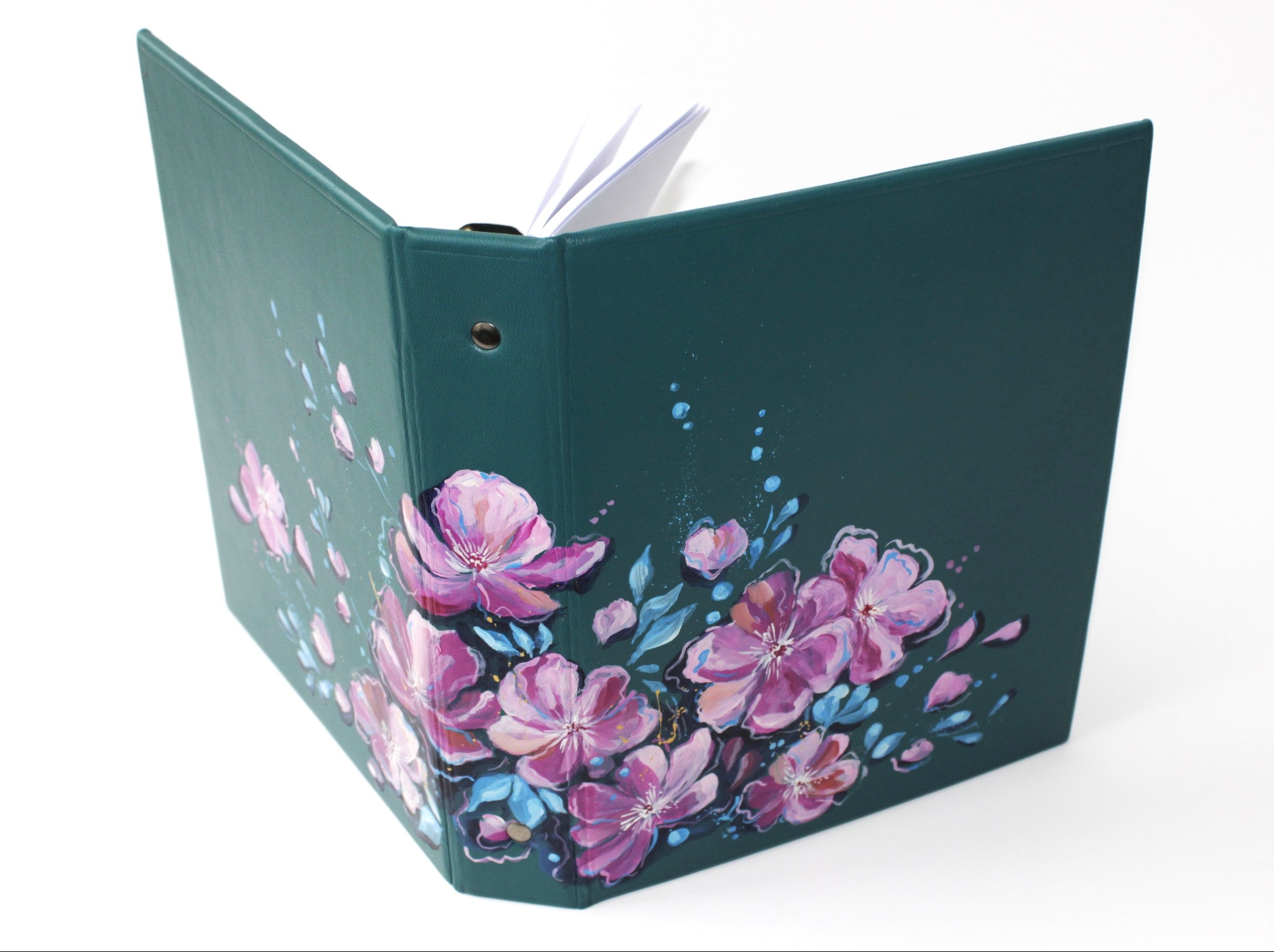 Otwarty, zielony segregator A5 z ręcznie malowanymi, różowymi kwiatami na okładce, widoczne białe kartki wewnątrz, na białym tle.