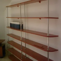 Regał na wymiar z półkami z drewna w kolorze wiśni, wsparty na metalowych, chromowanych rurach, z książkami na jednej z półek i ołówkiem na innej, ustawiony na tle białej ściany w pokoju na poddaszu.