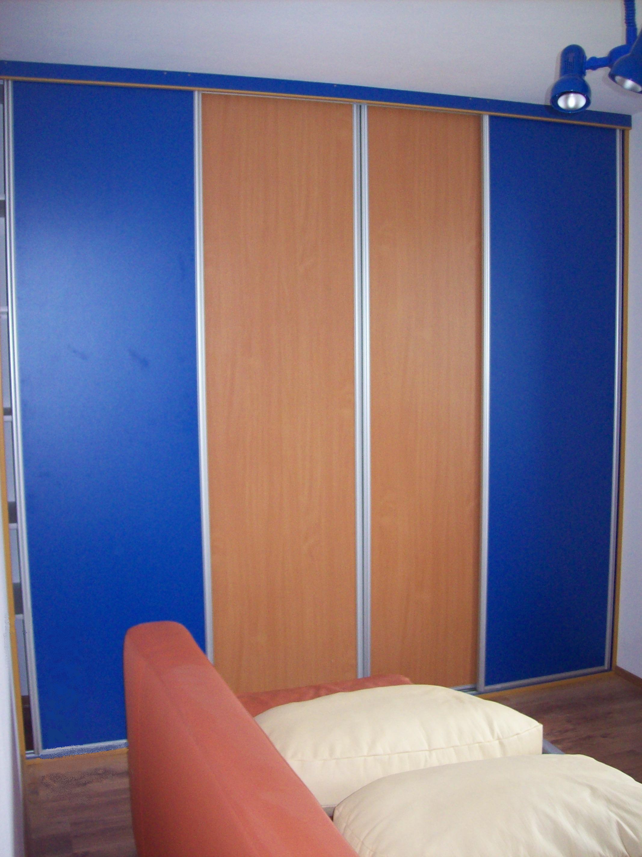 Zabudowa szafy przesuwnej z panelami w kolorze niebieskim i drewnopodobnym, widoczna z perspektywy pokoju z elementami sofy i poduszek.