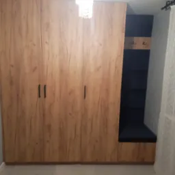 Trzydrzwiowa szafa na wymiar z drewnopodobnym wykończeniem, zintegrowana z wnęką z siedziskiem i wieszakami, oświetlona kryształowym żyrandolem.