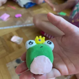 Dziecięca dłoń prezentuje figurkę żaby-króla wykonaną z masy polimerowej, z widocznymi elementami do modelowania w tle.