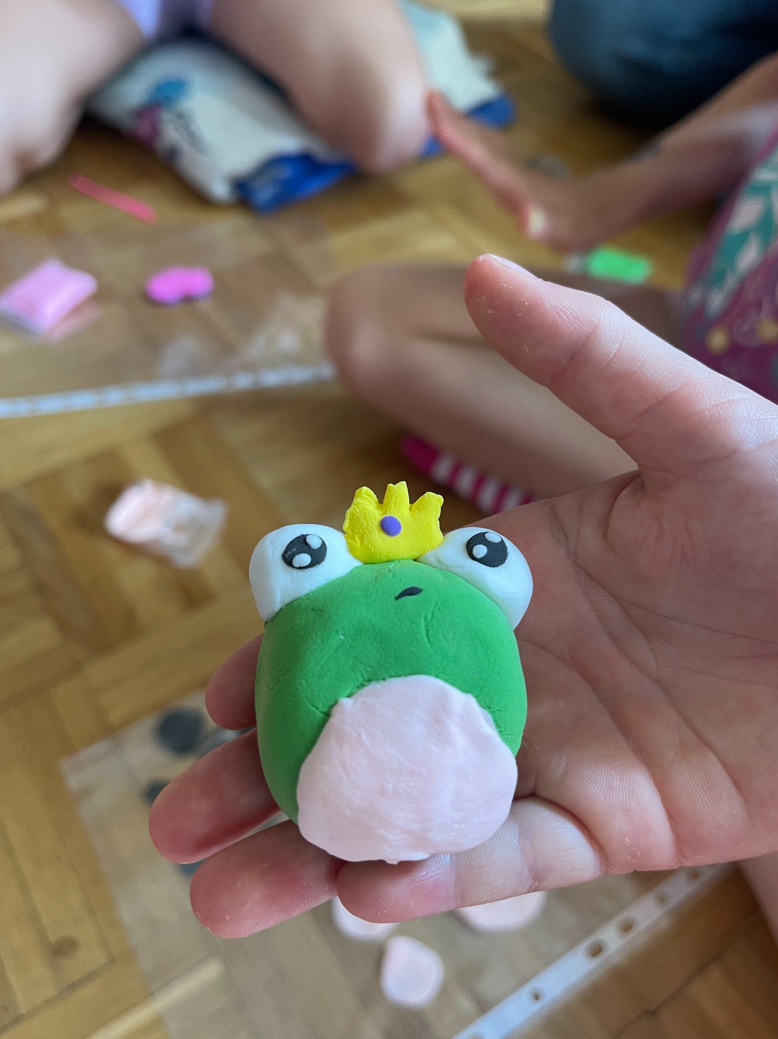 Dziecięca dłoń prezentuje figurkę żaby-króla wykonaną z masy polimerowej, z widocznymi elementami do modelowania w tle.