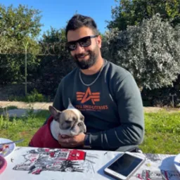 Mężczyzna w okularach przeciwsłonecznych siedzi na zewnątrz przy stole z psem rasy buldog francuski ubranym w czerwoną kurtkę, obok leży telefon komórkowy.