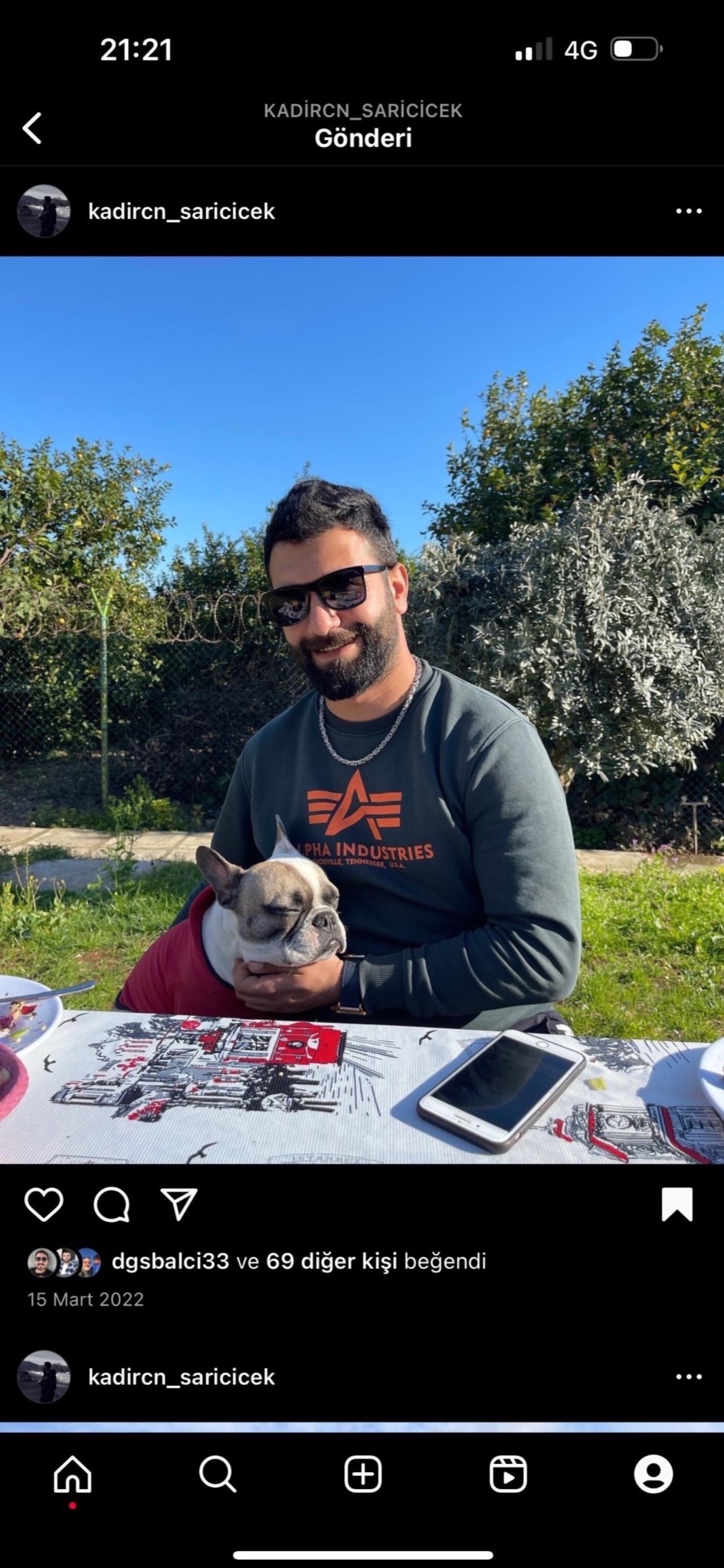 Mężczyzna w okularach przeciwsłonecznych siedzi na zewnątrz przy stole z psem rasy buldog francuski ubranym w czerwoną kurtkę, obok leży telefon komórkowy.