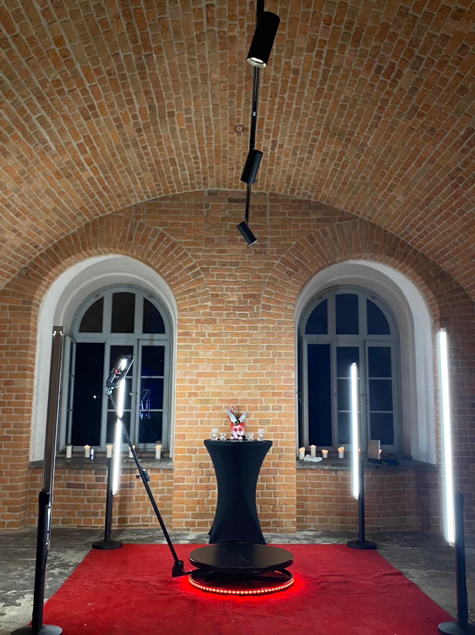 Okrągła platforma z czerwonym oświetleniem LED na czerwonym dywanie, otoczona lampami LED, ustawiona w ceglanym wnętrzu z łukowymi oknami i świecami.