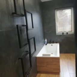 Szara łazienka z wanną obudowaną drewnem, dekoracyjnym czarnym grzejnikiem drabinkowym i roślinami w oknie.