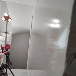 Usługi remontowo budowlane MAGBET - Świeżo nałożona, mokra gładź na ścianie w klatce schodowej, oświetlona lampą budowlaną Milwaukee, widoczny fragment schodów i sylwetka pracownika.