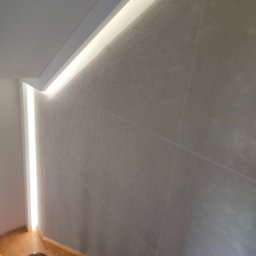Usługi remontowo budowlane MAGBET - Ściana wykończona gładzią strukturalną, oświetlona listwą LED wzdłuż sufitu, z widocznym fragmentem schodów z drewnianymi stopniami.