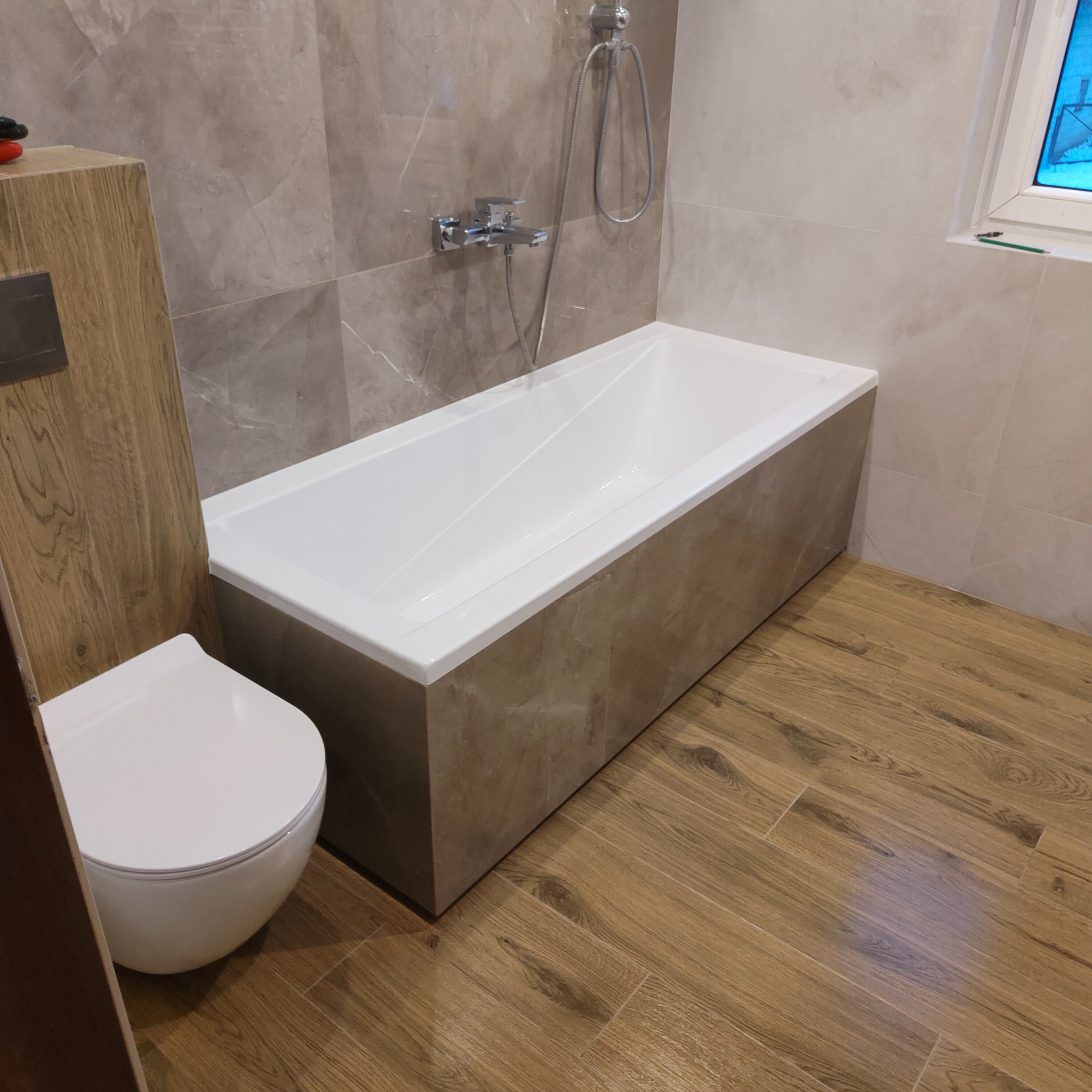 Nowoczesna łazienka z wanną obudowaną płytami imitującymi marmur, wiszącą toaletą i podłogą z paneli drewnopodobnych.