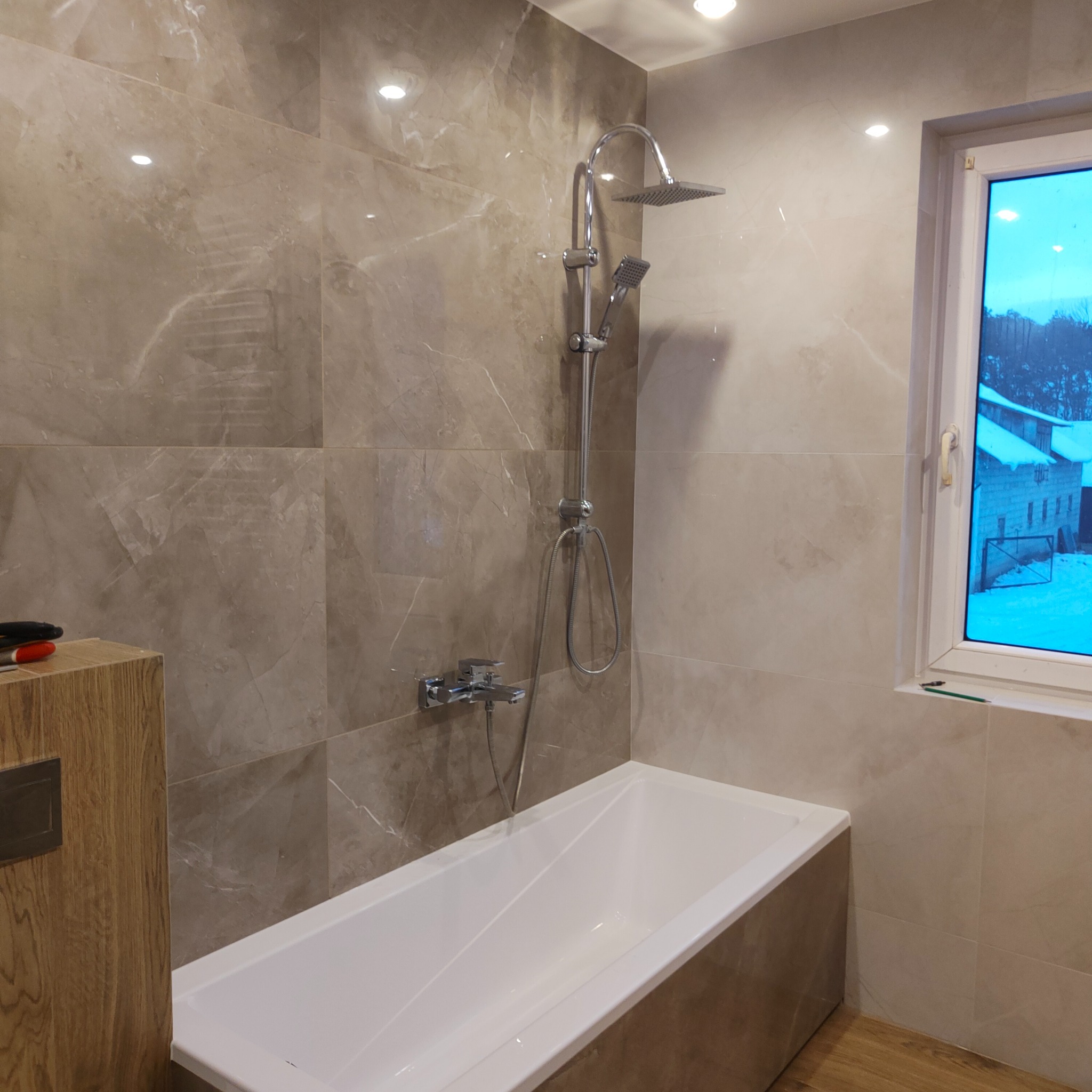 Nowoczesna łazienka z wanną, deszczownicą i marmurowymi płytkami w odcieniach beżu, widok na zasypany śniegiem krajobraz za oknem.