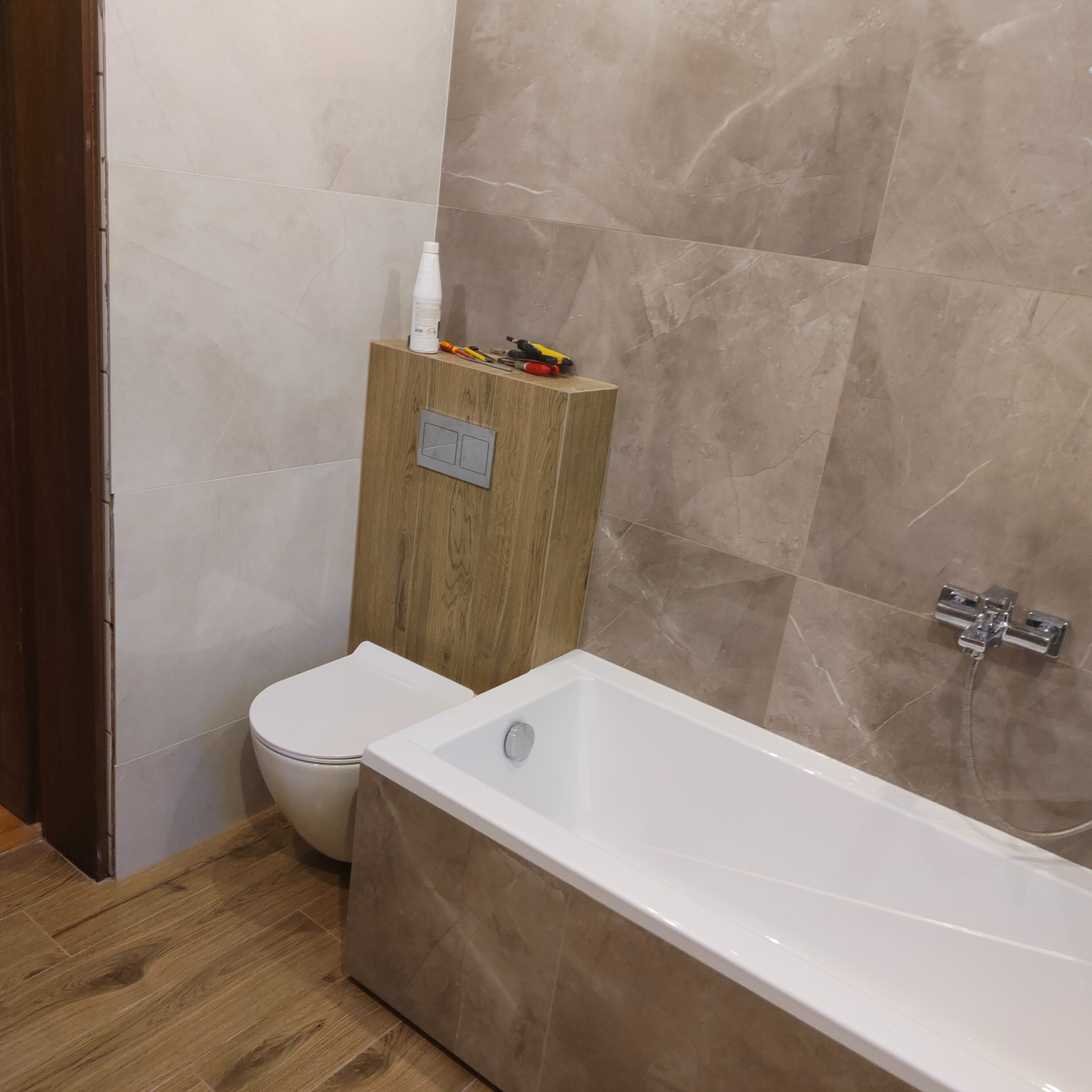 Wykończona łazienka z beżowymi płytkami imitującymi marmur, zabudowana toaleta z drewnianą obudową, biała wanna i drewnopodobna podłoga, na obudowie toalety leżą narzędzia i butelka.