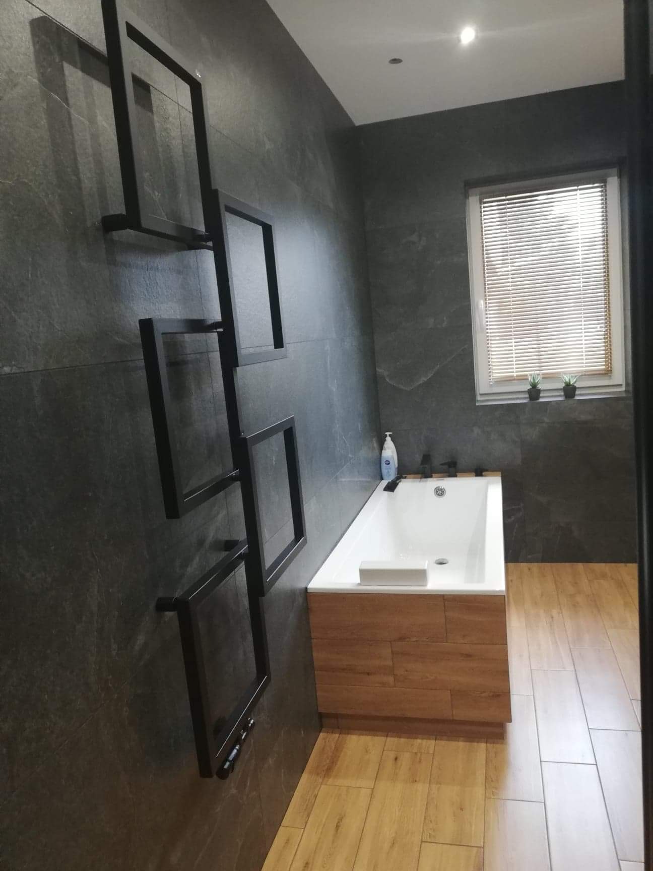 Szara łazienka z wanną obudowaną drewnem, dekoracyjnym czarnym grzejnikiem drabinkowym i roślinami w oknie.