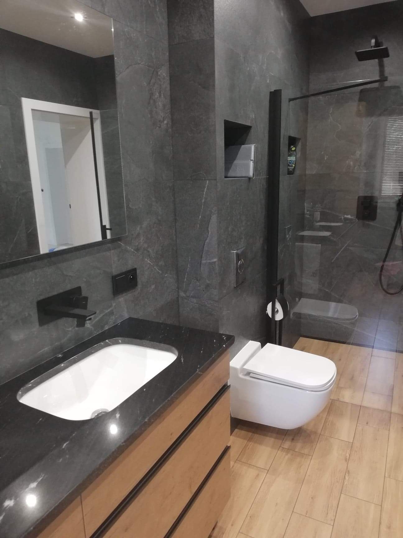 Nowoczesna łazienka z szarymi płytkami imitującymi kamień, drewnianą szafką pod umywalką z czarnym blatem, wiszącą toaletą i kabiną prysznicową typu walk-in z czarnymi profilami.