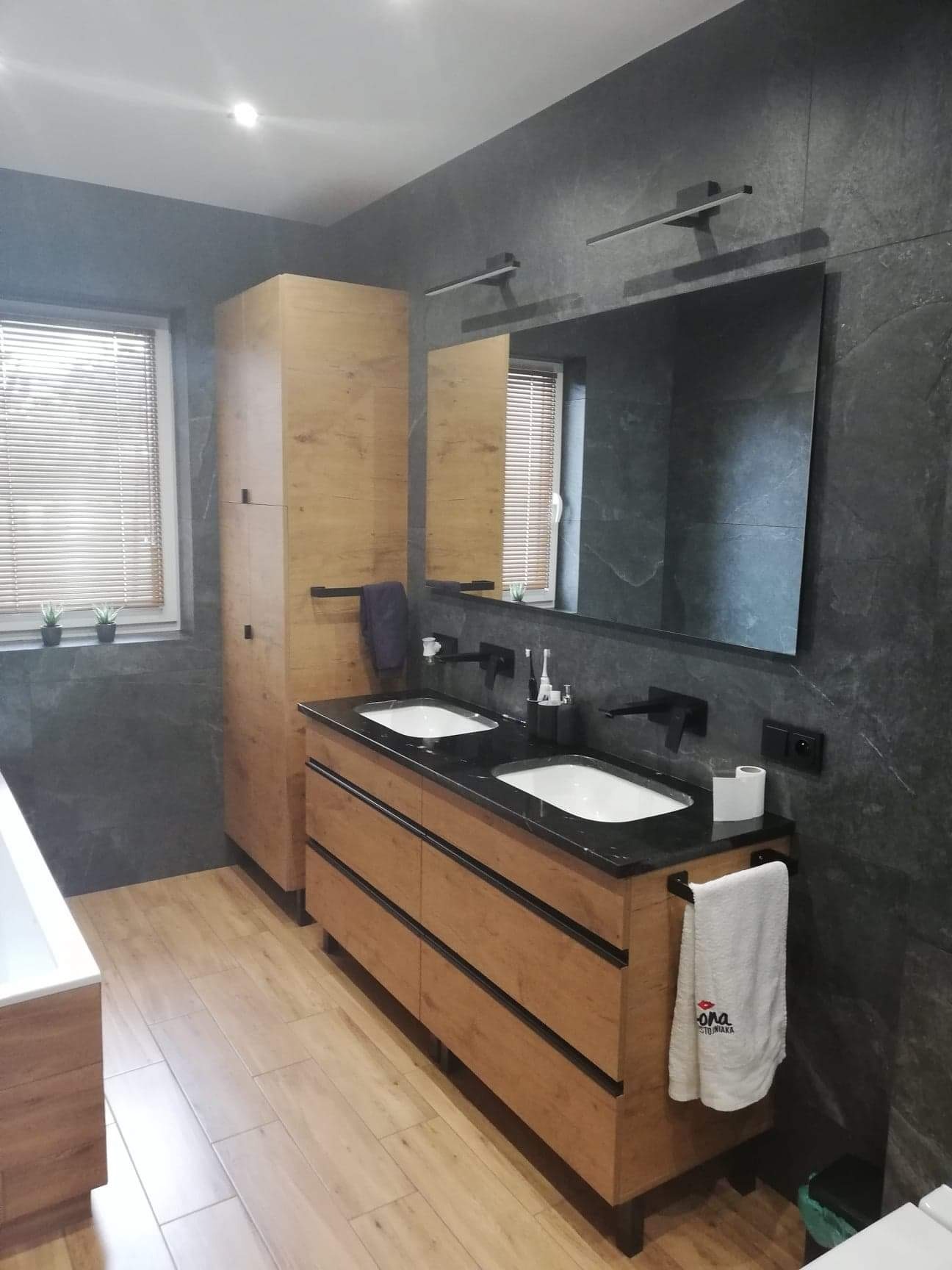 Nowoczesna łazienka z drewnianą szafką pod umywalki z czarnym blatem, szarą ścianą z efektem kamienia, dużym lustrem i drewnianą podłogą.