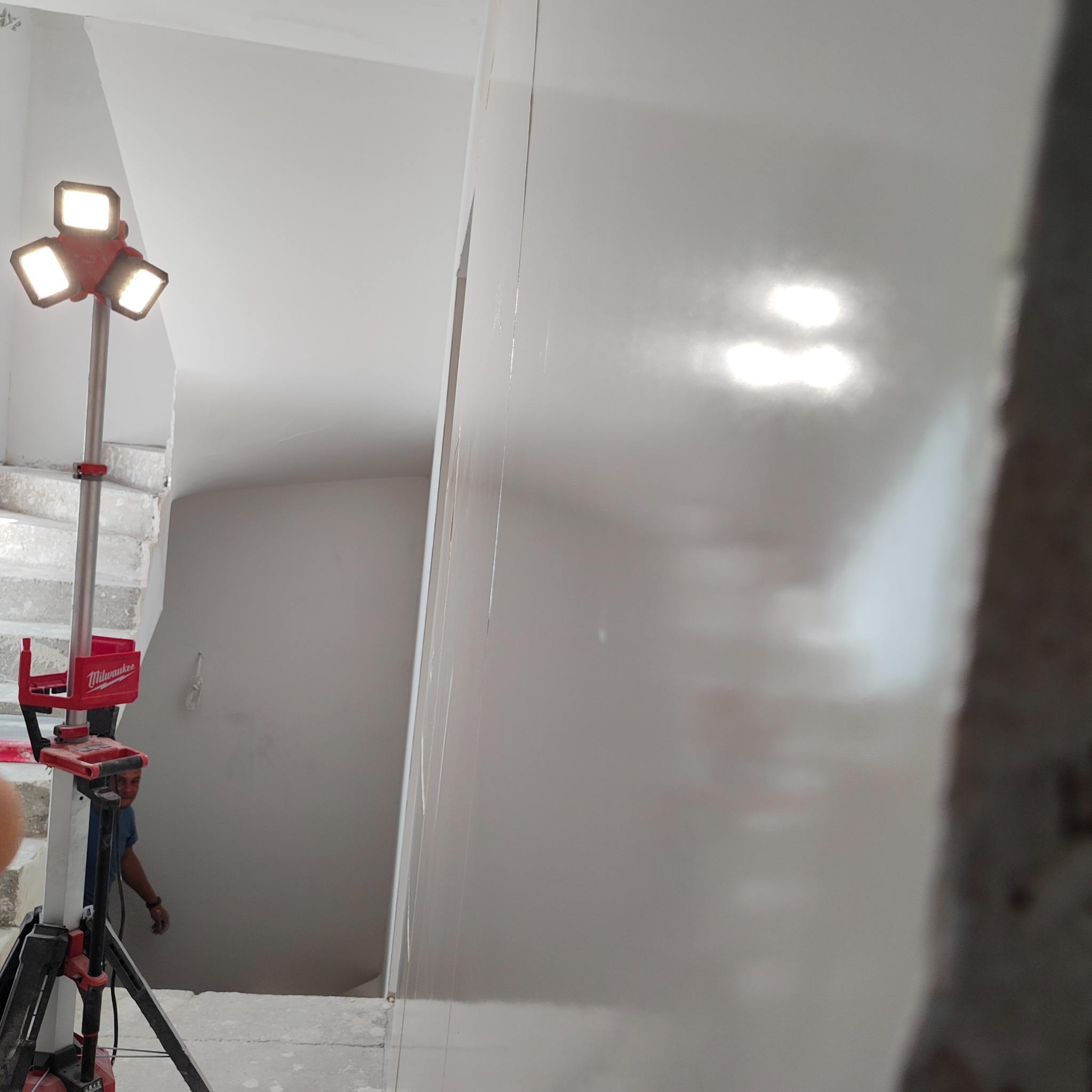 Świeżo nałożona, mokra gładź na ścianie w klatce schodowej, oświetlona lampą budowlaną Milwaukee, widoczny fragment schodów i sylwetka pracownika.