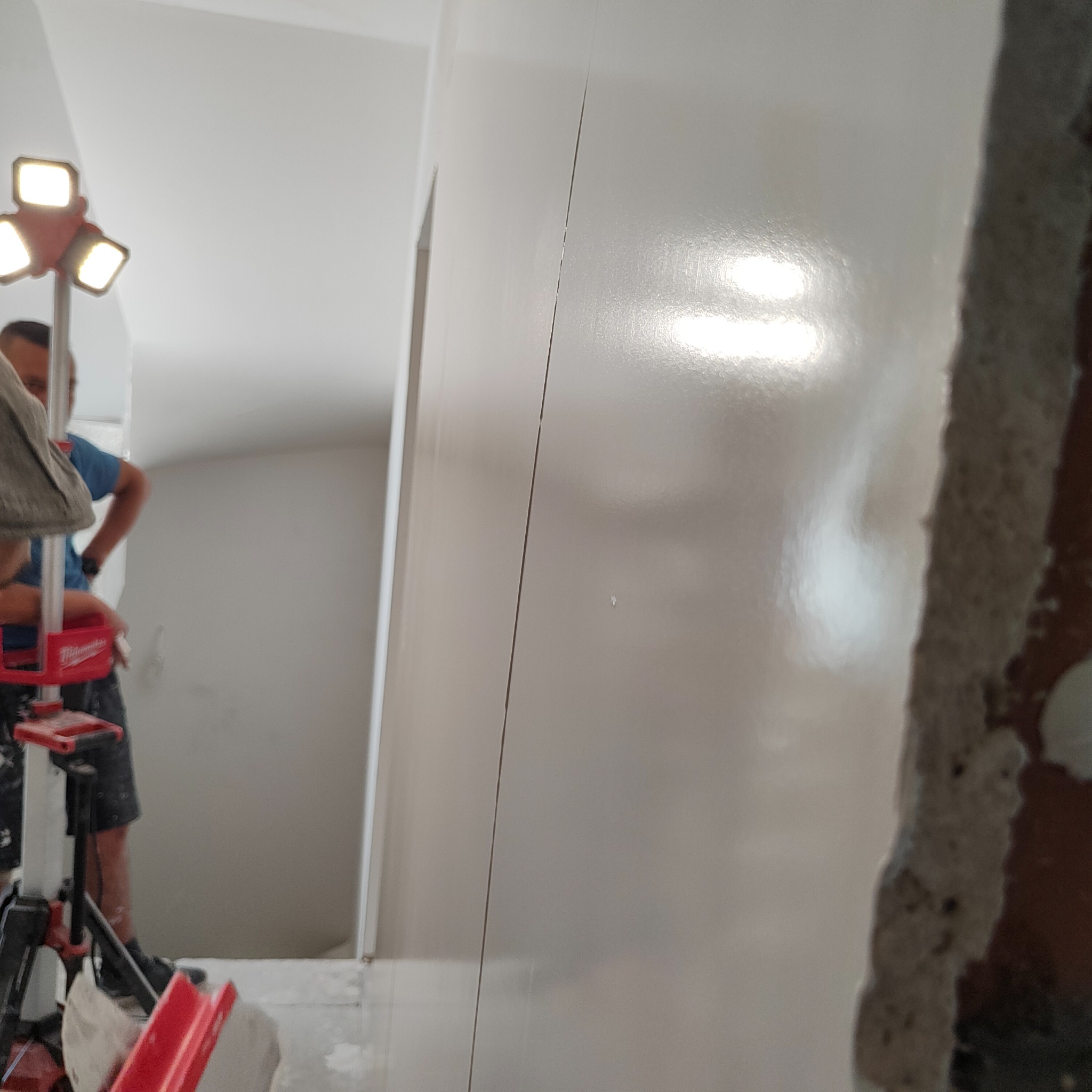 Świeżo położona gładź na ścianie, widoczna spoina, oświetlenie robocze Milwaukee, fragment muru w tle.