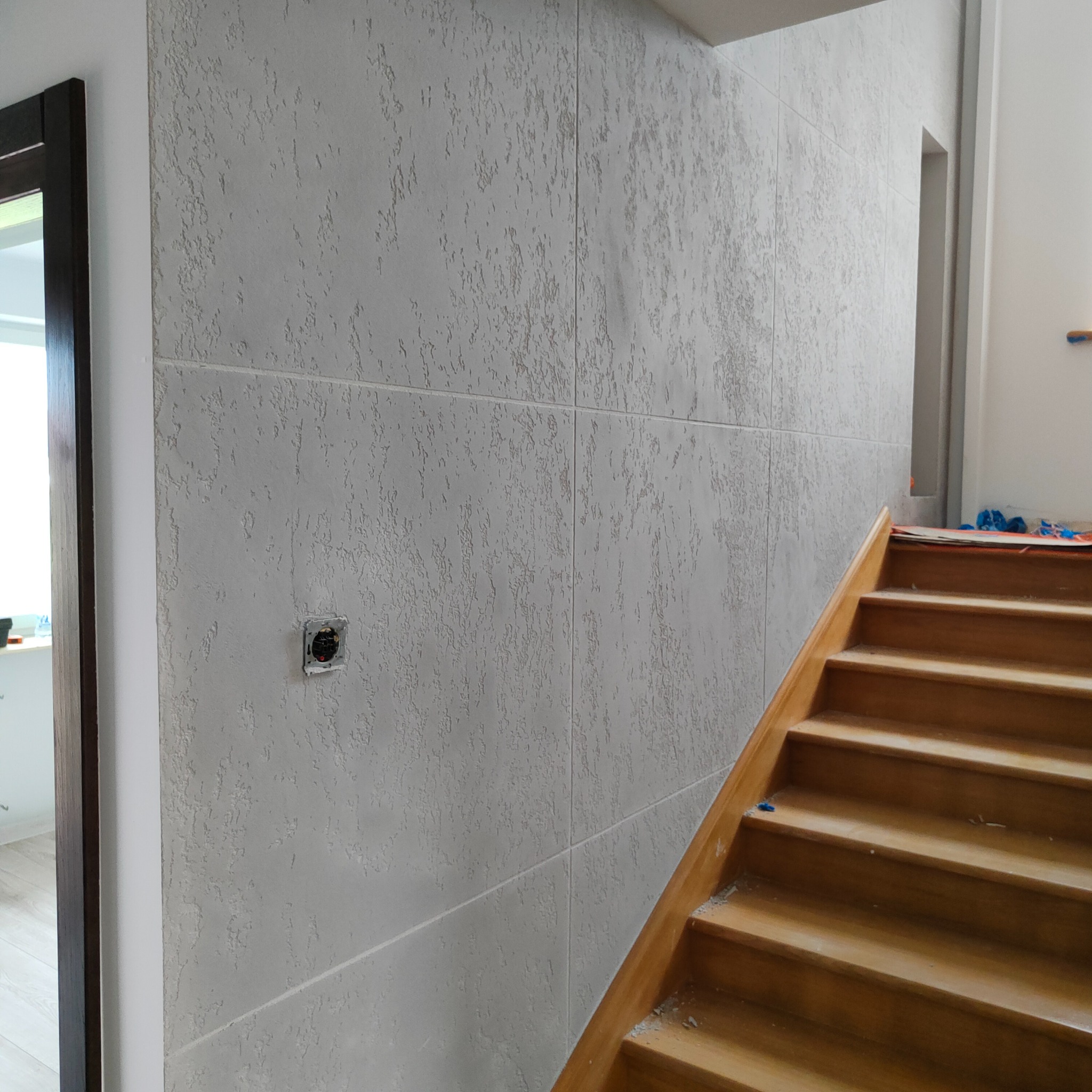 Ściana wykończona szarą gładzią dekoracyjną z widocznym gniazdkiem elektrycznym i fragmentem drewnianych schodów w trakcie remontu.