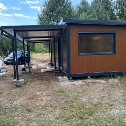PAWILONY LL Łukasz Leśnowolski - Nowoczesny pawilon z płyt warstwowych z zadaszeniem, widoczny na tle lasu i błękitnego nieba. Na pierwszym planie rozłożone materiały budowlane i samochód.