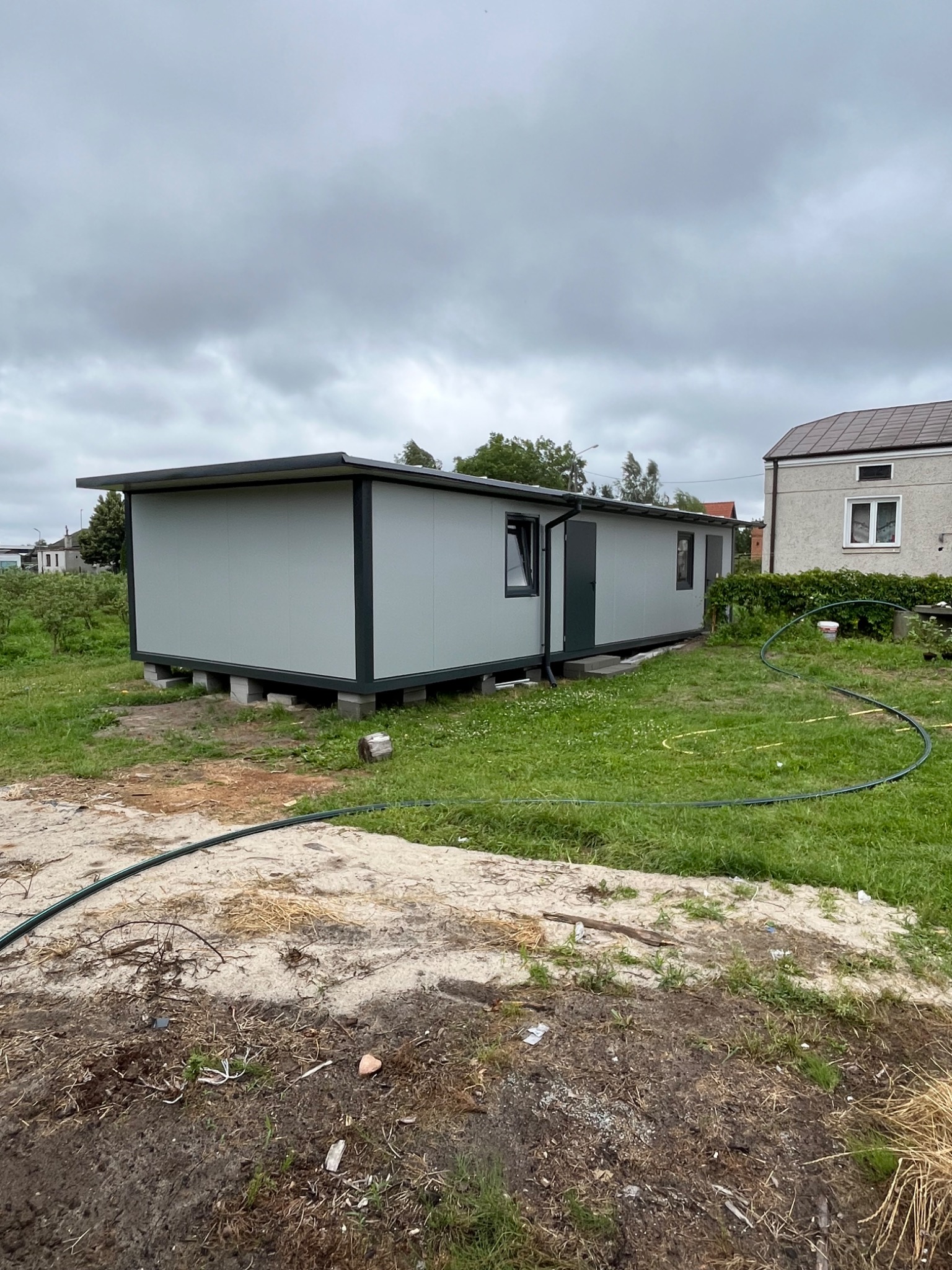 Szary budynek z płyt warstwowych z ciemnymi obramowaniami okien i drzwi, umieszczony na betonowych blokach na trawiastym terenie.