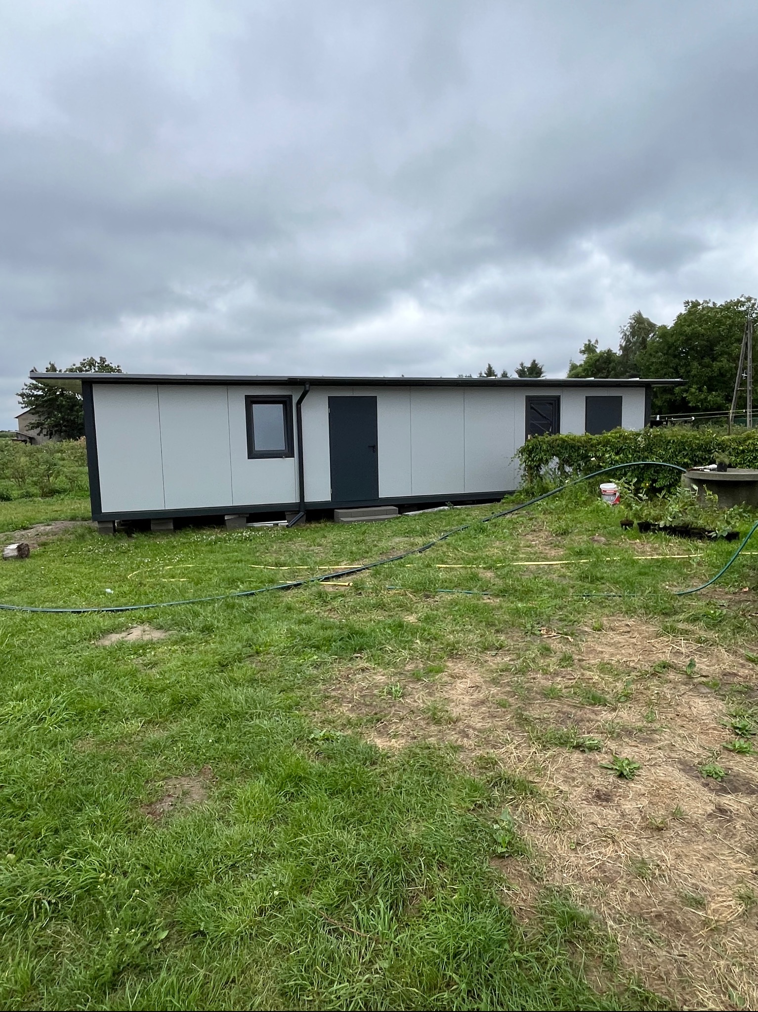 Długi, szary pawilon z płyt warstwowych z ciemnymi drzwiami i oknami, ustawiony na trawie pod pochmurnym niebem. Wokół leżą węże ogrodowe.