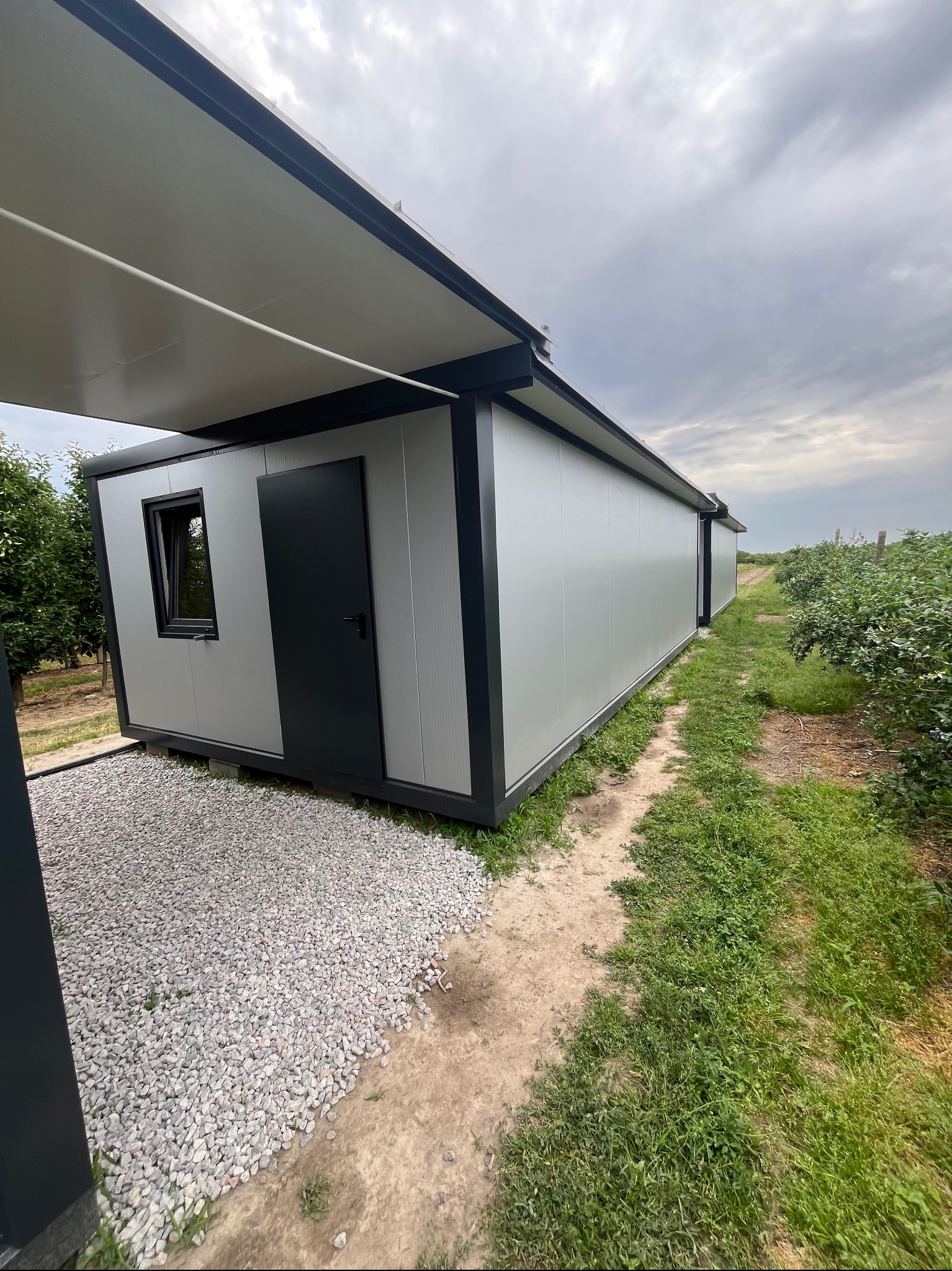 Nowoczesny pawilon z płyt warstwowych w otoczeniu zieleni, widok z zewnątrz. Szara elewacja, czarne detale, żwirowe podłoże. Letnie popołudnie.