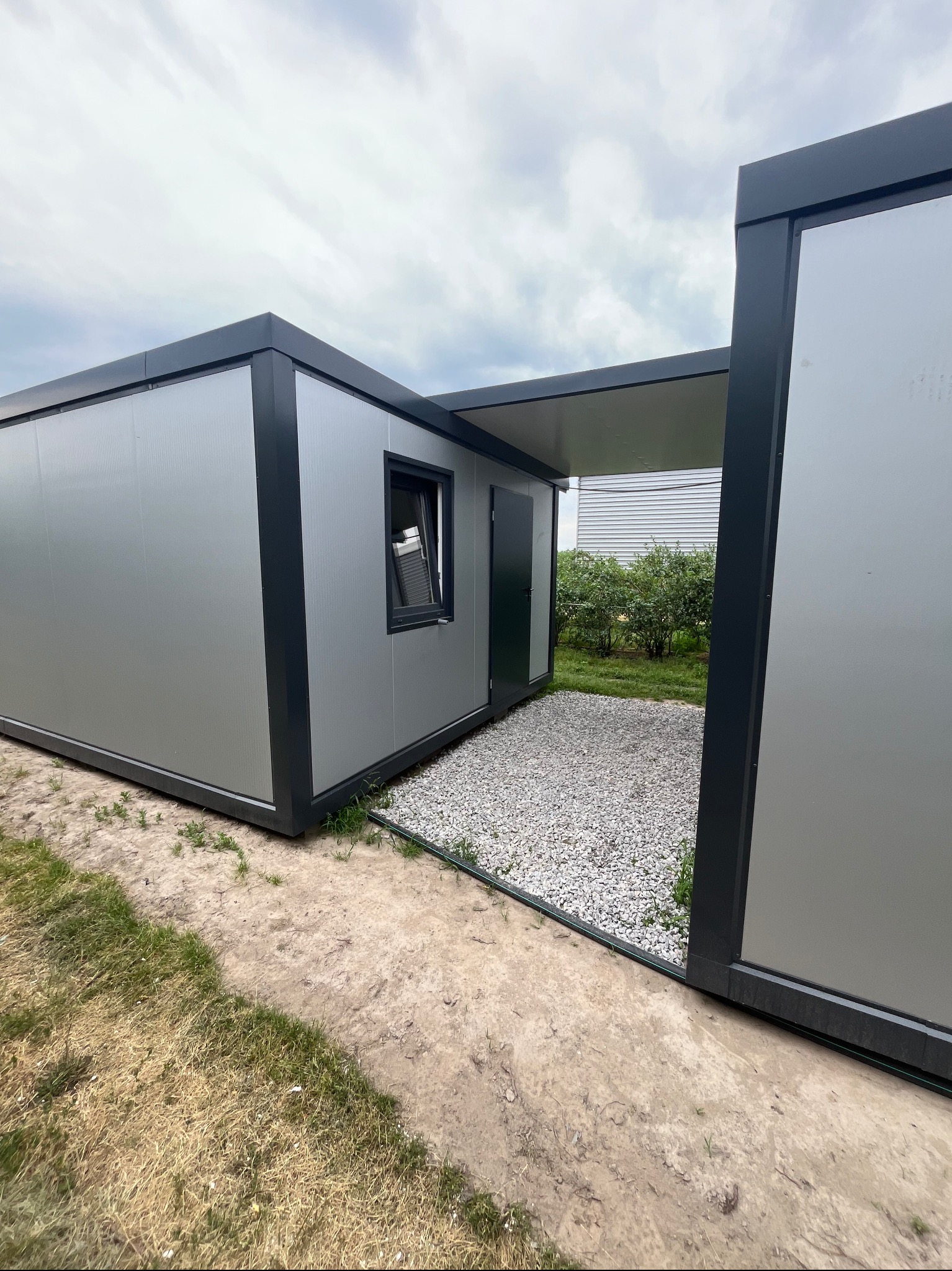 Nowoczesny pawilon z płyt warstwowych w kolorze szarym z czarnymi obramowaniami, z widocznym oknem i drzwiami, otoczony żwirem i trawą, pod pochmurnym niebem.