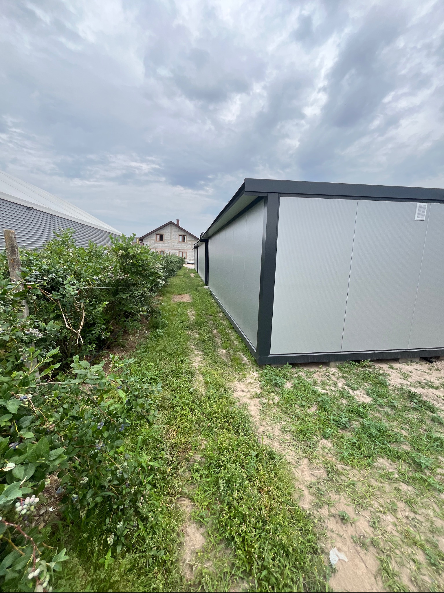 Kontener z płyt warstwowych w szarym kolorze, z czarnymi obramowaniami, umiejscowiony na zewnątrz, w tle zabudowania i zachmurzone niebo.