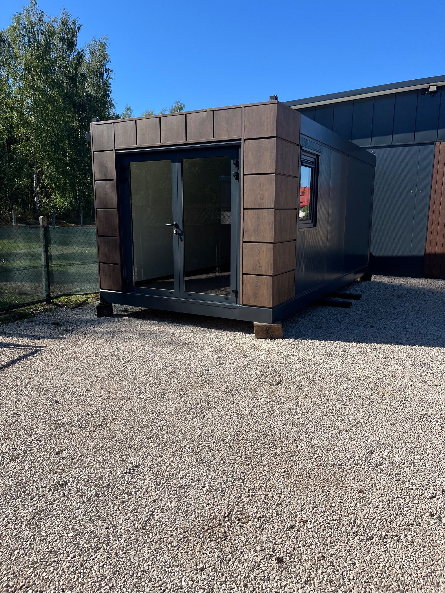 Gotowy modułowy budynek z płyt warstwowych, z przeszklonym wejściem, ustawiony na żwirowym podłożu. Widoczne elementy wykończeniowe imitujące drewno.