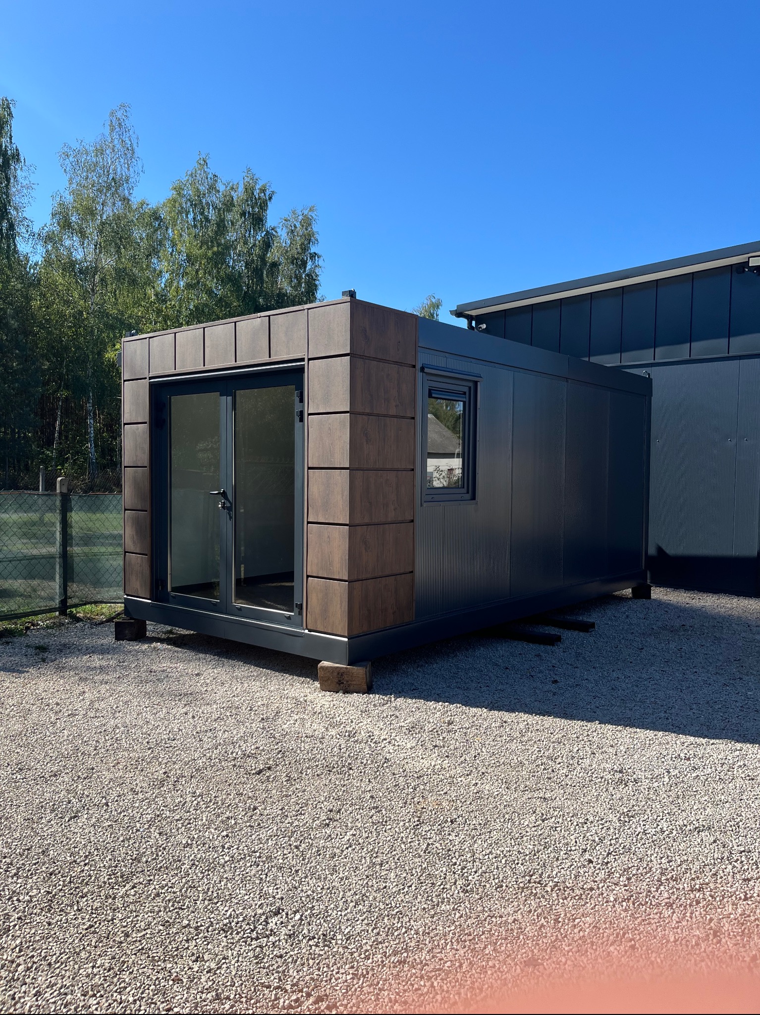 Gotowy modułowy budynek z płyt warstwowych, z drewnopodobną elewacją i przeszklonymi drzwiami, ustawiony na żwirowym podłożu w słoneczny dzień.
