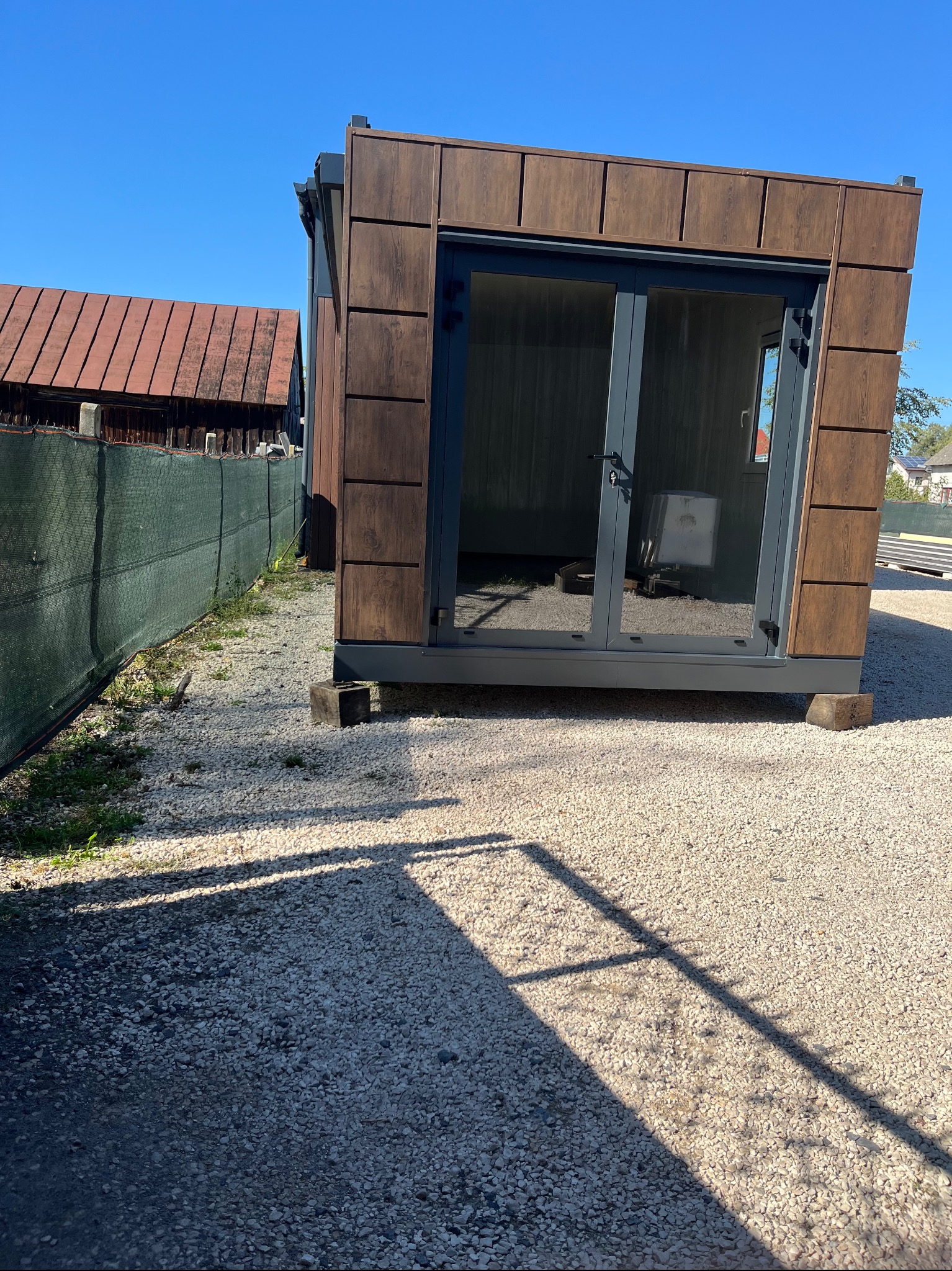 Gotowy modułowy budynek z płyt warstwowych o drewnopodobnej fakturze, z dużymi szklanymi drzwiami, ustawiony na żwirowym podłożu w słoneczny dzień.