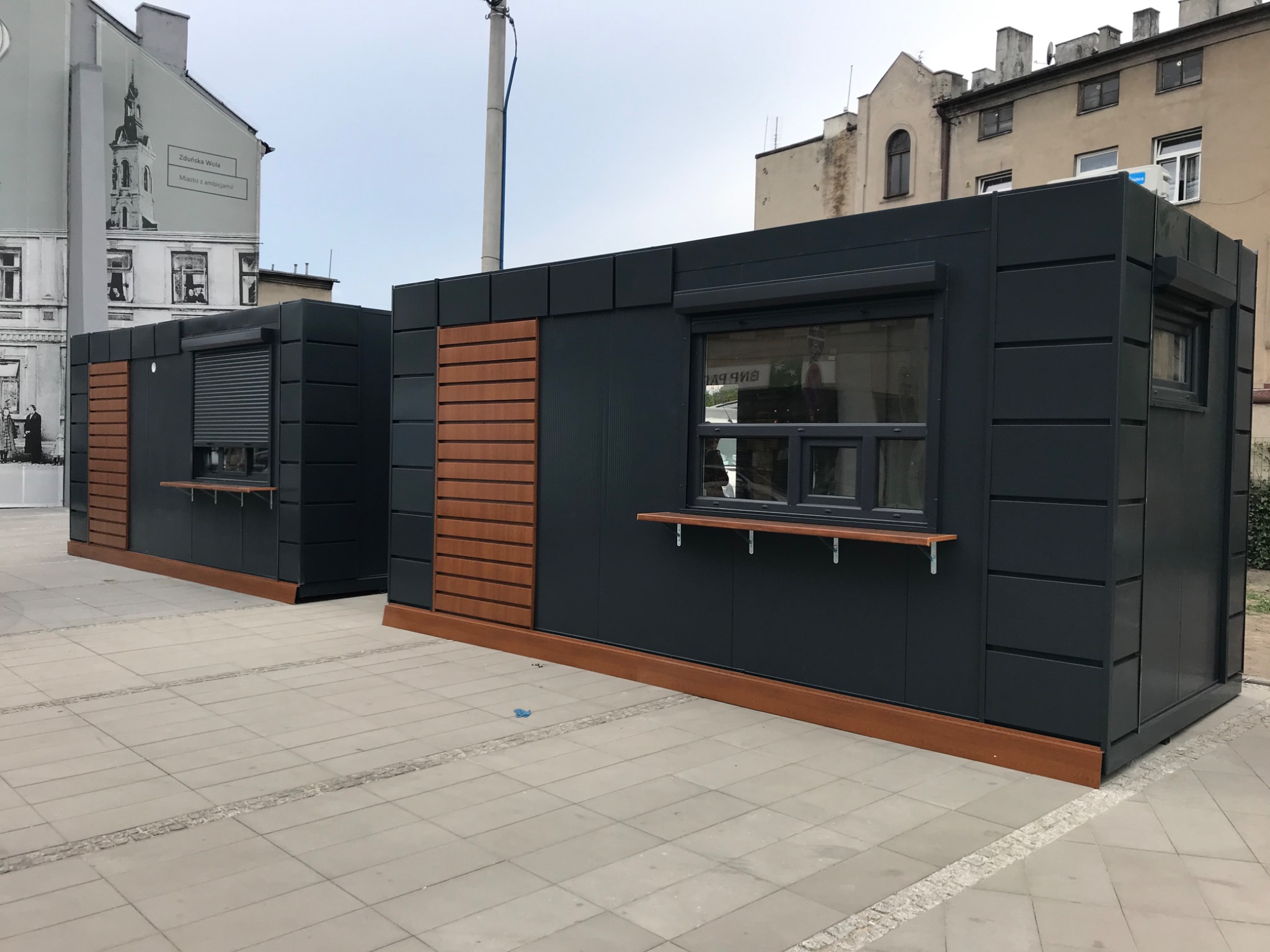 Dwa pawilony handlowe z płyt warstwowych w kolorze grafitowym, z drewnianymi elementami dekoracyjnymi, na tle miejskiej zabudowy. Jeden z pawilonów ma zasłonięte okno.